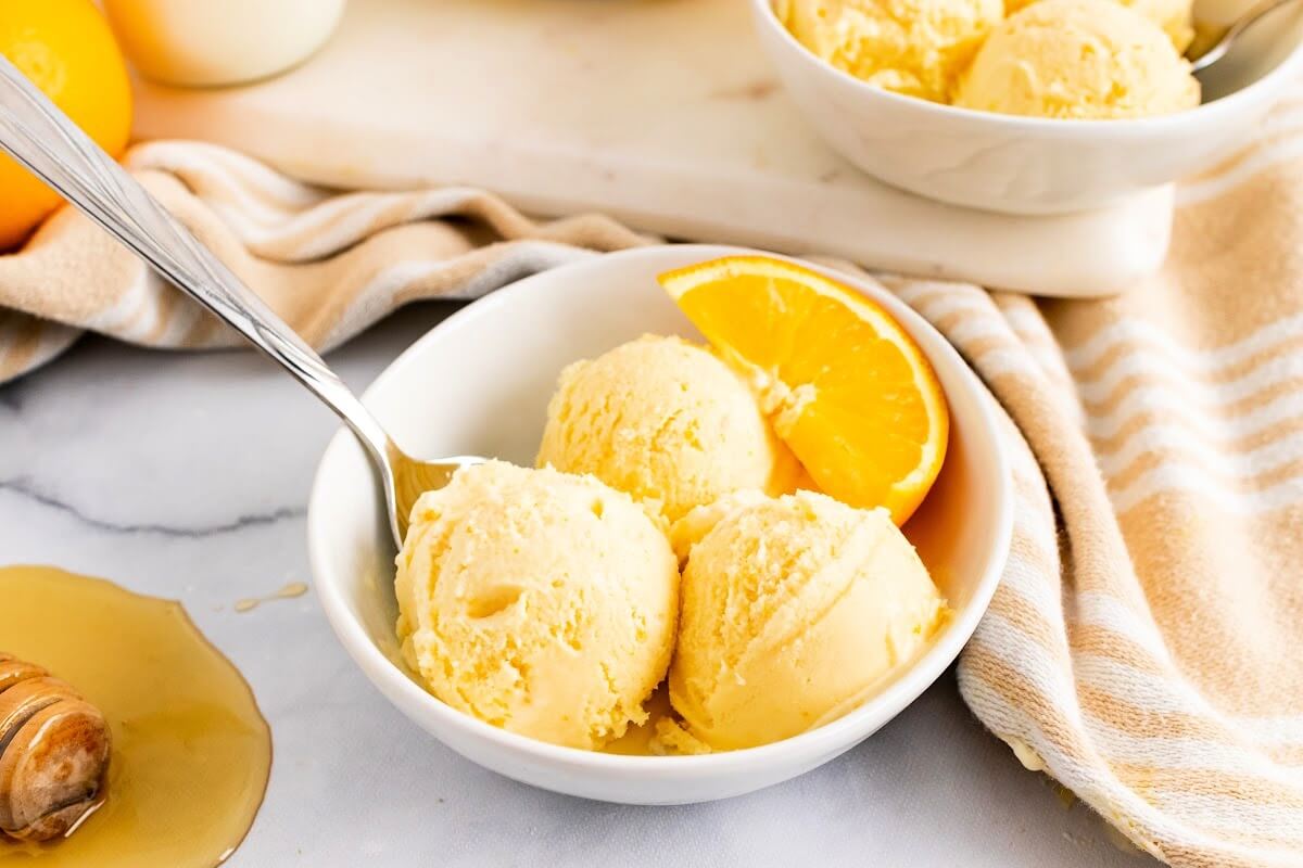 Bowl with scoops of orange ice cream, a spoon and fresh orange slices and honey spilled next to it.