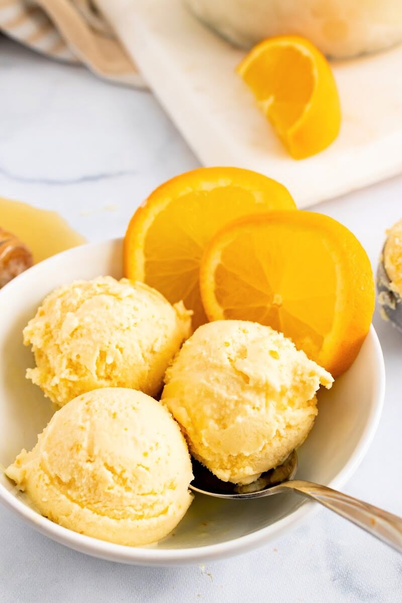 Bowl with scoops of orange ice cream, a spoon and fresh orange slices.