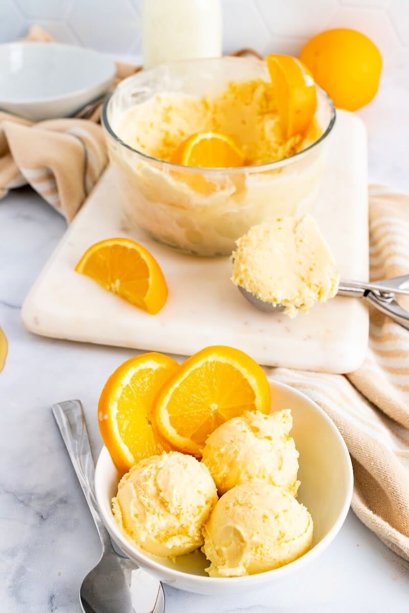 Bowls with scoops of orange ice cream, a spoon and fresh orange slices sitting next to a large bowl full of ice cream and orange slices sitting on top of a marble cutting board with an ice cream scooper filled with ice cream.