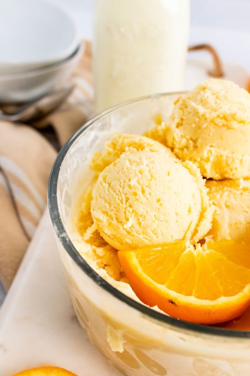 Glass bowl with scoops of orange ice cream and fresh orange slices next to a bottle of milk and empty bowls.
