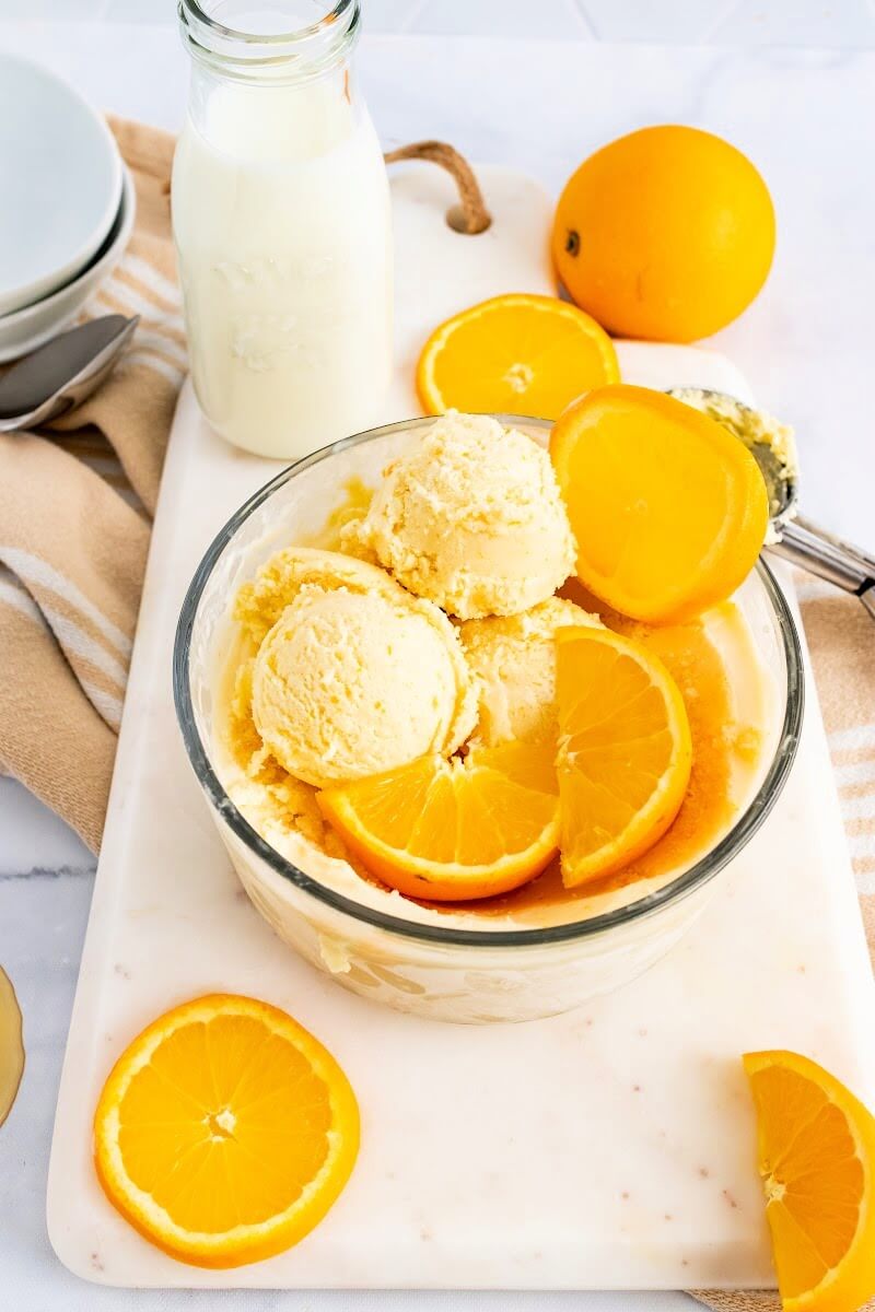 Large bowl full of scoops of orange ice cream and fresh orange slices sitting on top of a marble cutting board with an ice cream scooper, a bottle of milk, empty bowls and a cloth kitchen towel next to it.
