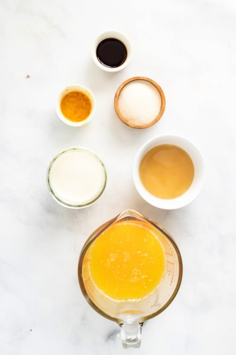 Glass measuring cup filled with orange juice, small bowl with honey, small bowl with cream, small bowl with vanilla extract. small bowl with orange zest and small bowl with arrowroot powder.