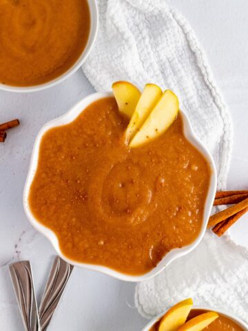 Three bowls filled with homemade applesauce with slices of apple, one dusted with ground cinnamon, sitting next to a towel, spoons and cinnamon sticks.