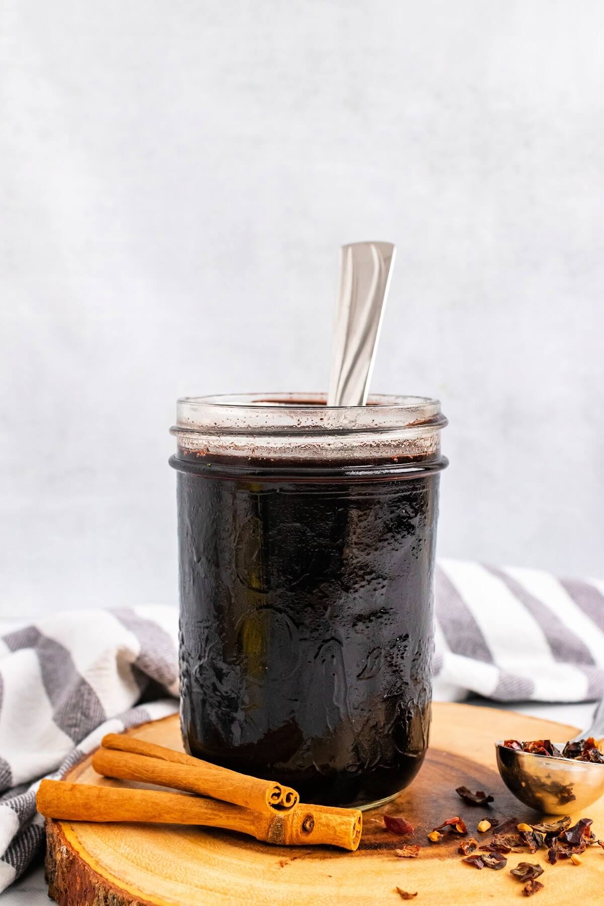 Mason jar filled with dark purple syrup with a spoon in it, sitting on top of a wood tree slab circle cutting board, next to cinnamon sticks and a spoon full of dried rose hips with a cloth kitchen towel behind it.