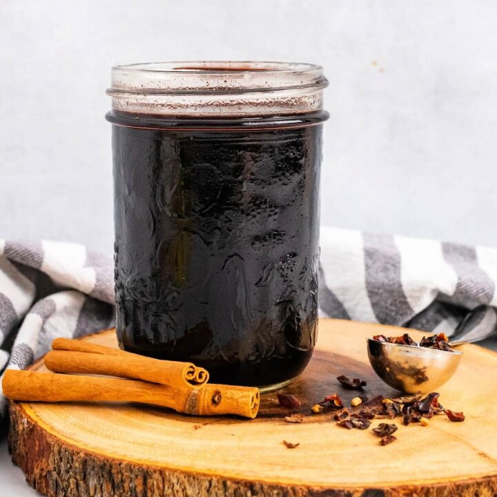 Mason jar filled with dark purple syrup, sitting on top of a wood tree slab circle cutting board, next to cinnamon sticks and a spoon full of dried rose hips with a cloth kitchen towel behind it.