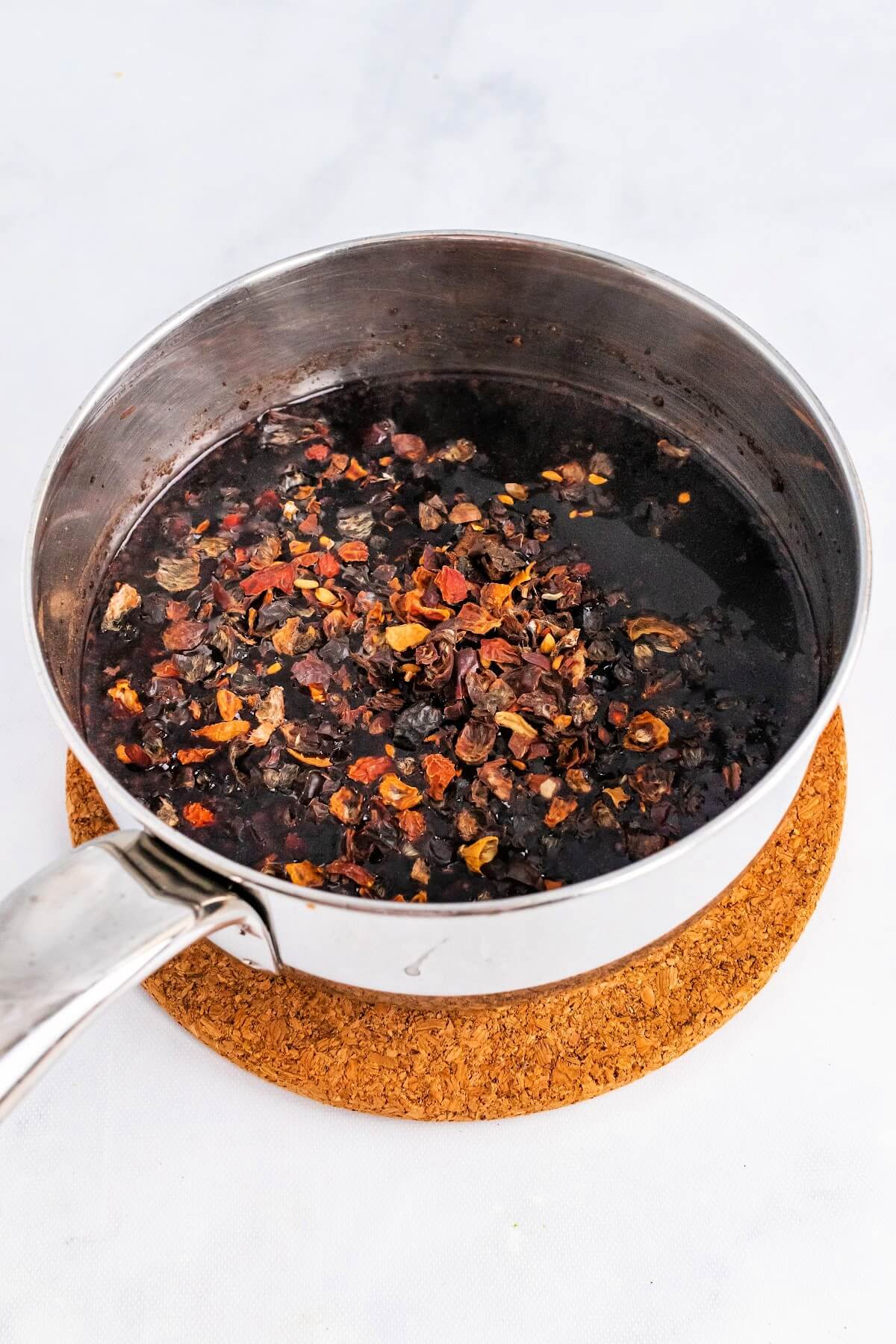 Saucepan filled with a dark syrup and dried rose hips floating at the top, sitting on top of a cork trivet.