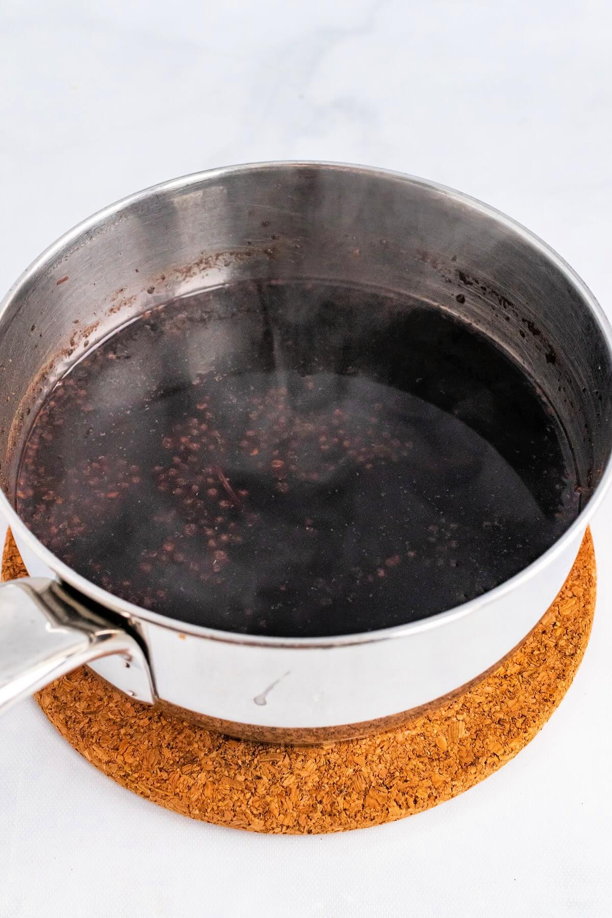 Saucepan filled with a steaming hot dark purple liquid and cooked elderberries floating at the top, sitting on top of a cork trivet.