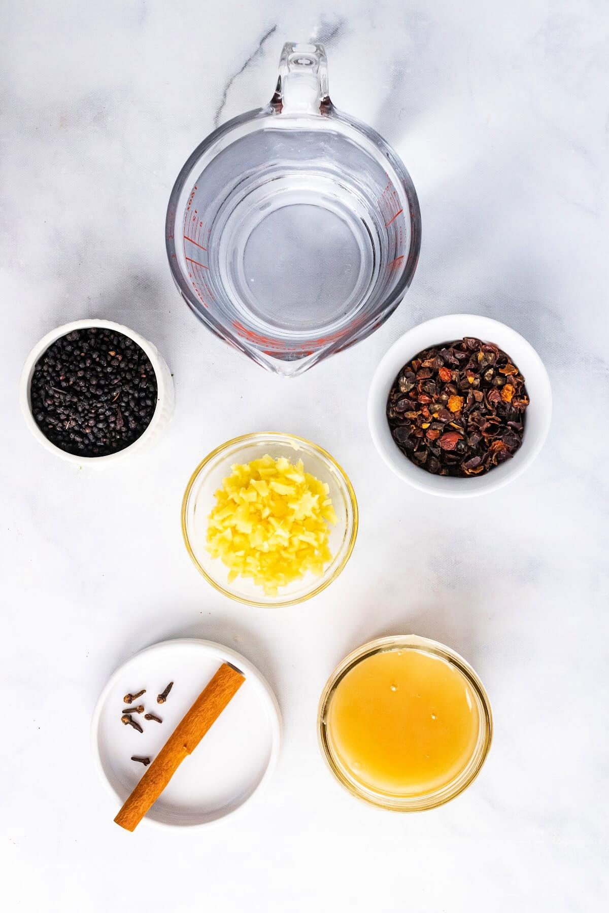 Overhead view of a glass measuring cup full of water, a small bowl filled with dried elderberries, a small bowl filled with dried rose hips, a small bowl filled with chopped fresh ginger, a small bowl filled with honey and a small dish with a cinnamon stick and whole cloves.