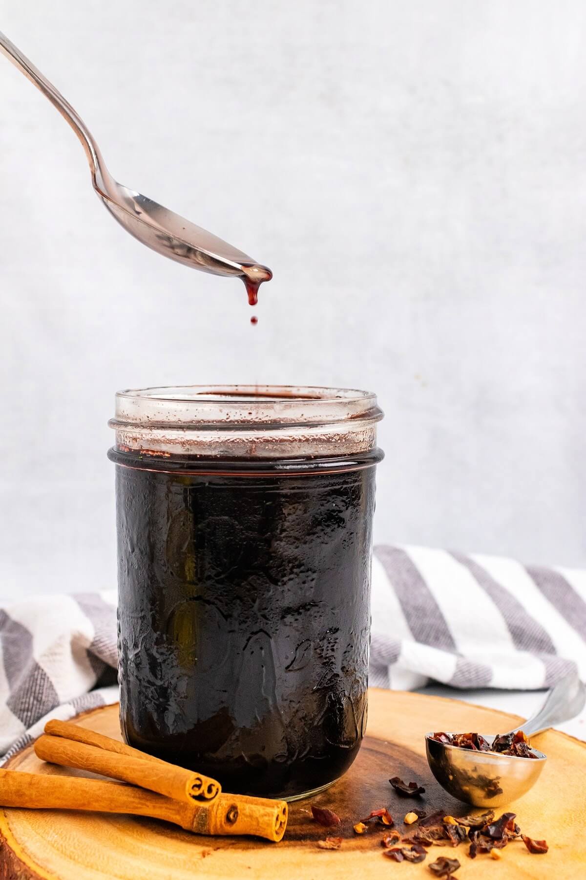 Mason jar filled with dark purple syrup, sitting on top of a wood tree slab circle cutting board, next to cinnamon sticks and a spoon full of dried rose hips with a cloth kitchen towel behind it and a spoon above the mason jar dripping syrup.