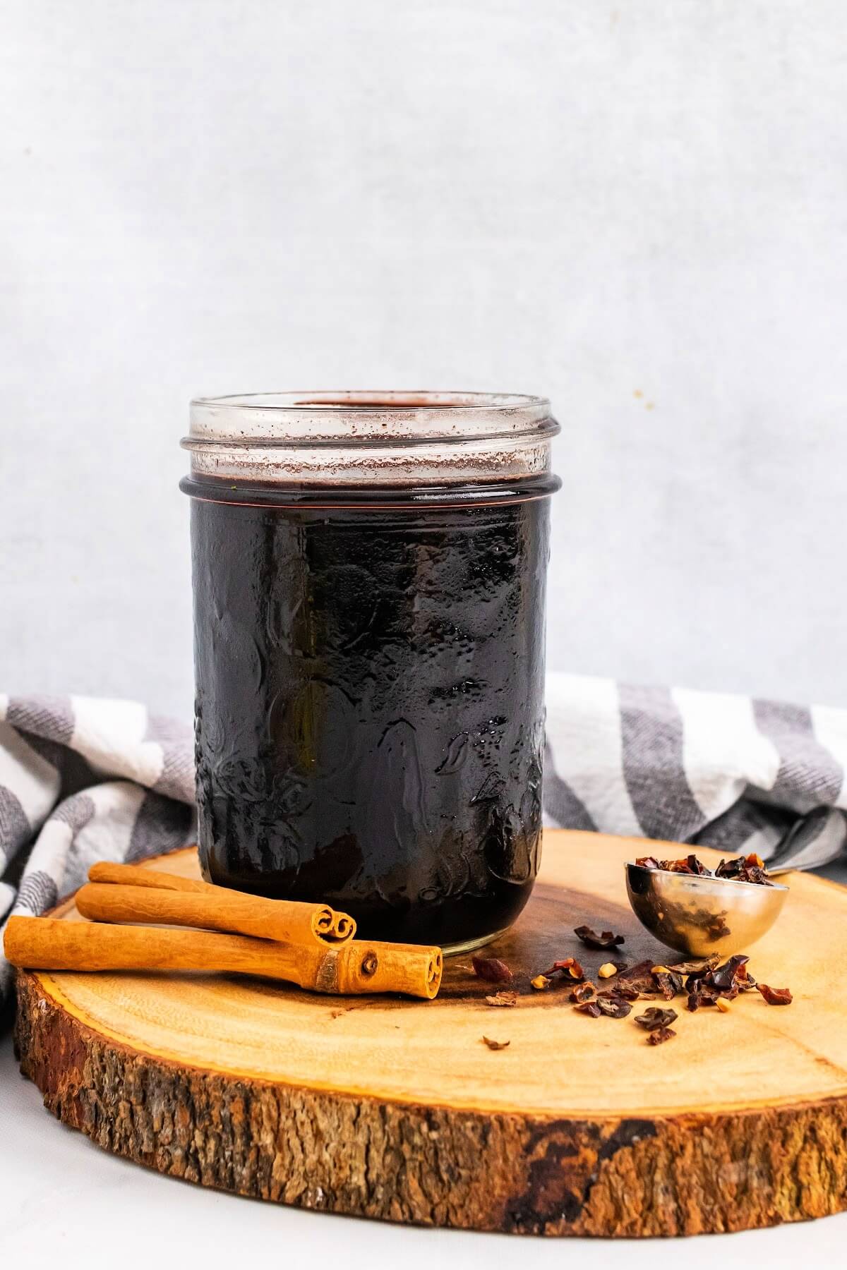 Mason jar filled with dark purple syrup, sitting on top of a wood tree slab circle cutting board, next to cinnamon sticks and a spoon full of dried rose hips with a cloth kitchen towel behind it.