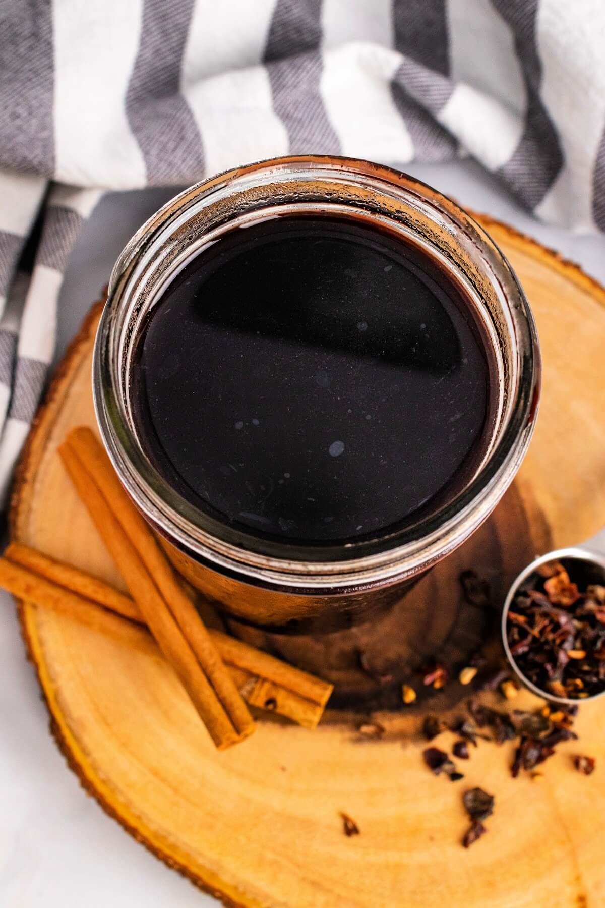 Overhead view of a mason jar filled with dark purple syrup, sitting on top of a wood tree slab circle cutting board, next to cinnamon sticks and a spoon full of dried rose hips with a cloth kitchen towel behind it.