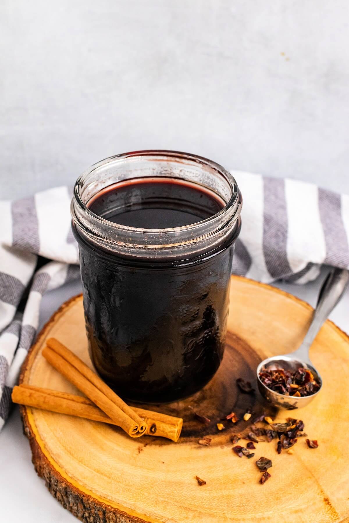Mason jar filled with dark purple syrup, sitting on top of a wood tree slab circle cutting board, next to cinnamon sticks and a spoon full of dried rose hips with a cloth kitchen towel behind it.