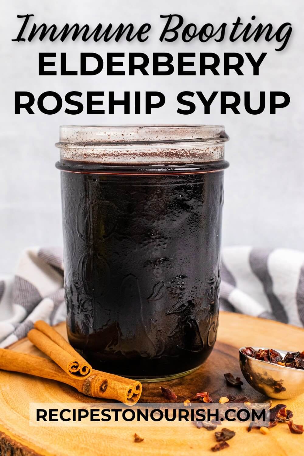 Mason jar filled with dark purple syrup, sitting on top of a wood tree slab circle cutting board, next to cinnamon sticks and a spoon full of dried rose hips with a cloth kitchen towel behind it and text that says Immune Boosting Elderberry Rosehip Syrup.