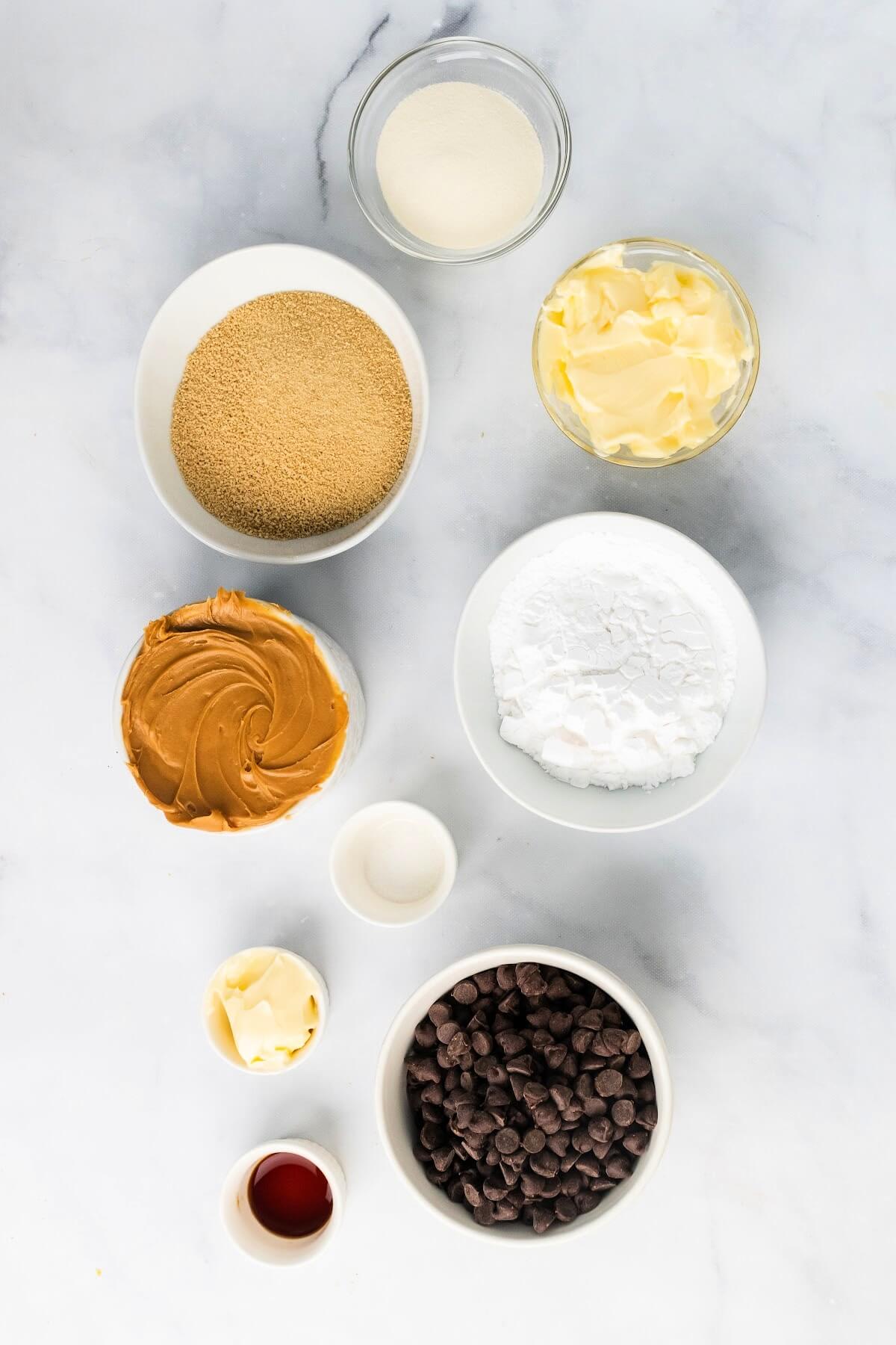 An overhead shot of a small bowl with collagen, a small bowl with butter, a small bowl with maple sugar, a small bowl with peanut butter, a small bowl with arrowroot powder, a mini bowl of vanilla extract, a bowl of chocolate chips, a mini bowl off salt and a mini bowl butter.