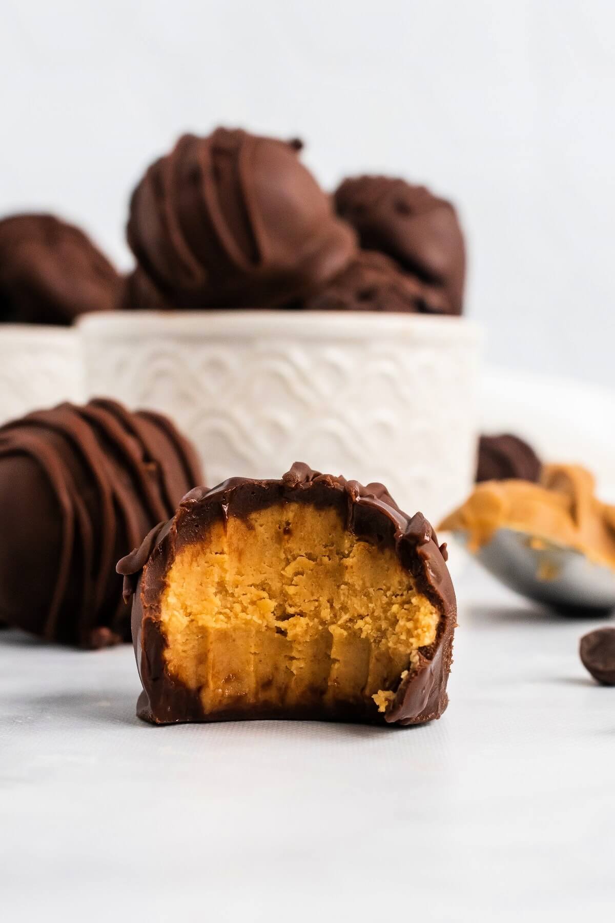 A homemade chocolate covered peanut butter truffle with a bite taken out of it sitting in front of another whole chocolate covered truffle, next to a spoon full of peanut butter and a bowl behind them full of chocolate covered truffles.