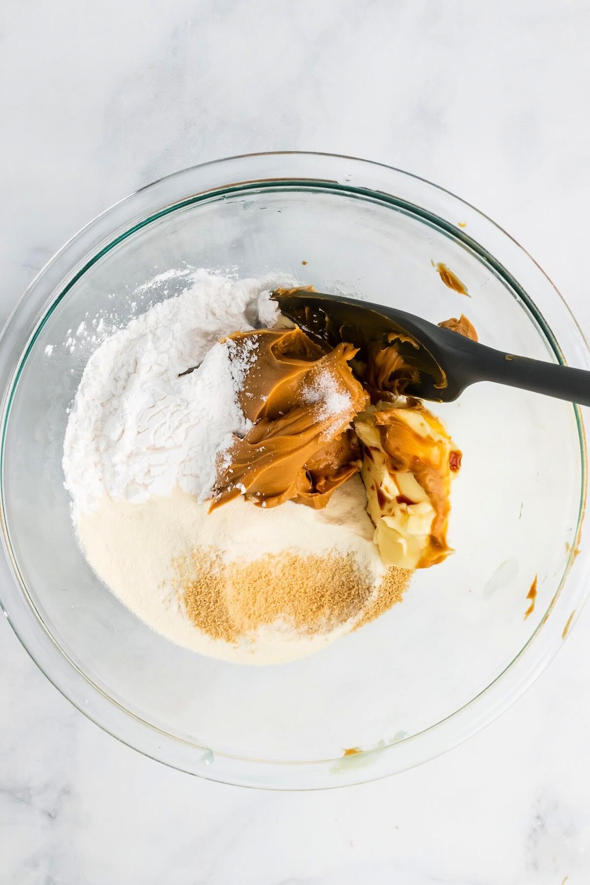 A glass mixing bowl filled with peanut butter, butter, vanilla, arrowroot powder, collagen, maple sugar and a spatula about to stir the mixture.