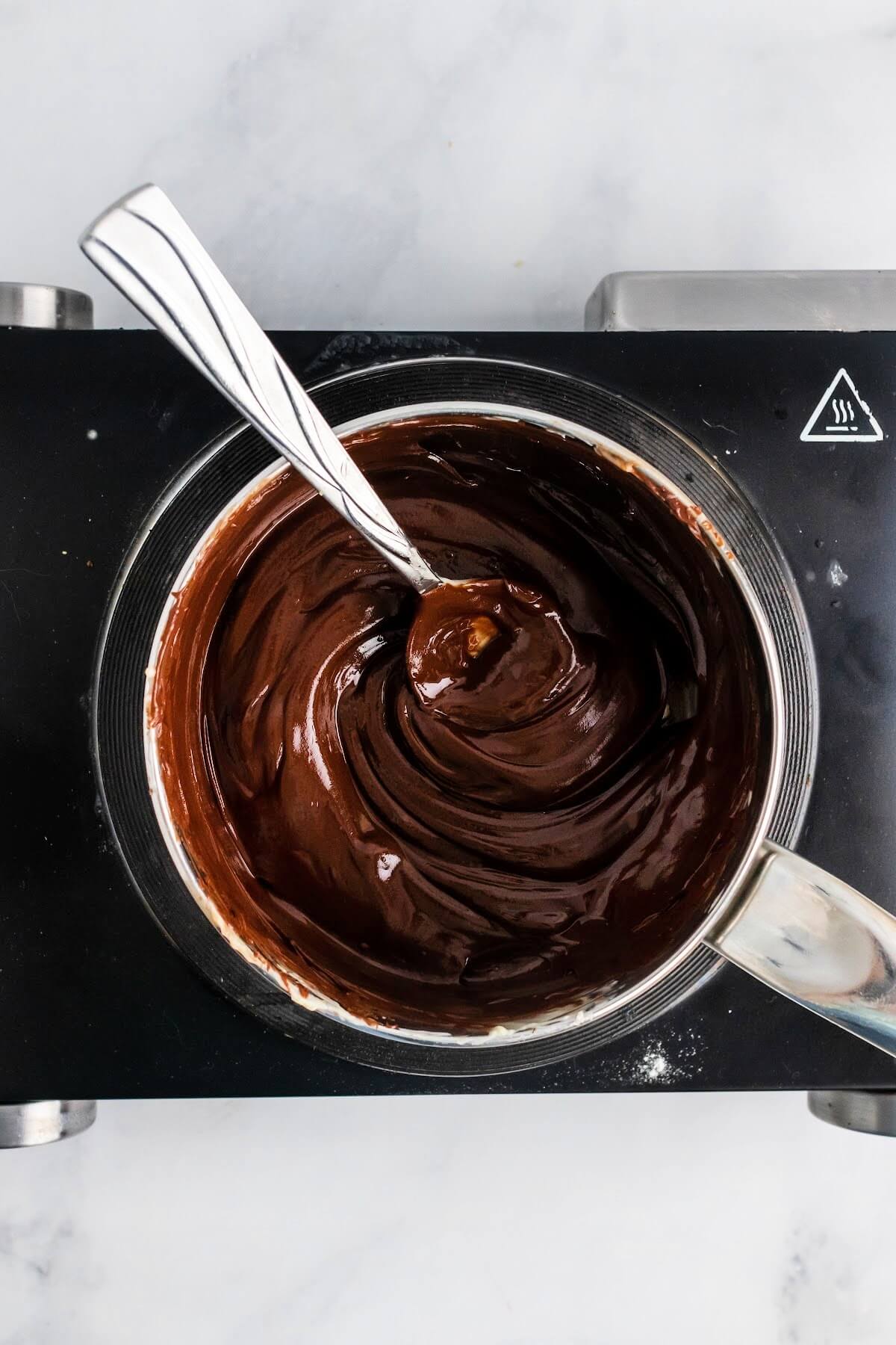 Saucepan with melted chocolate and a spoon stirring it sitting on top of an electric stovetop.