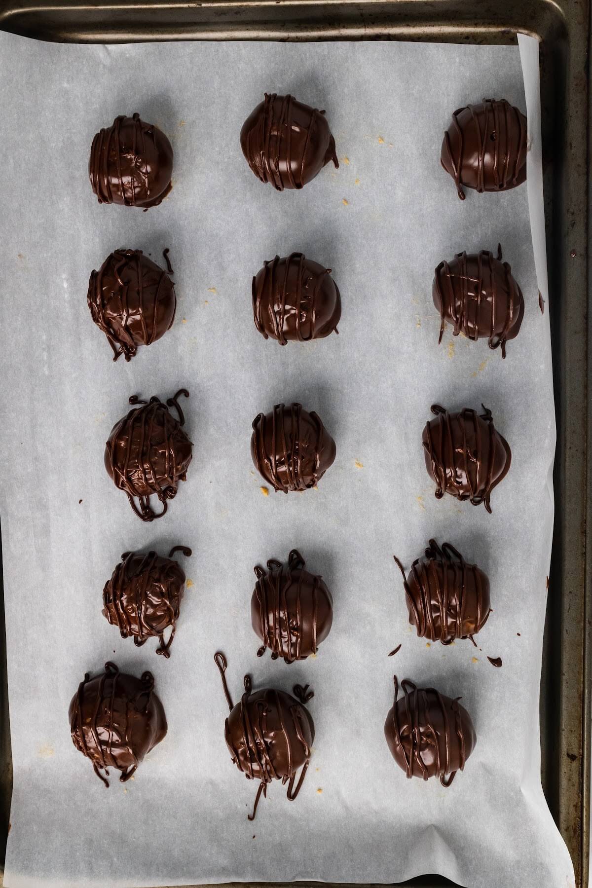 A baking sheet lined with parchment paper with 15 freshly coated chocolate truffles with drizzled chocolate over the tops of the truffles.