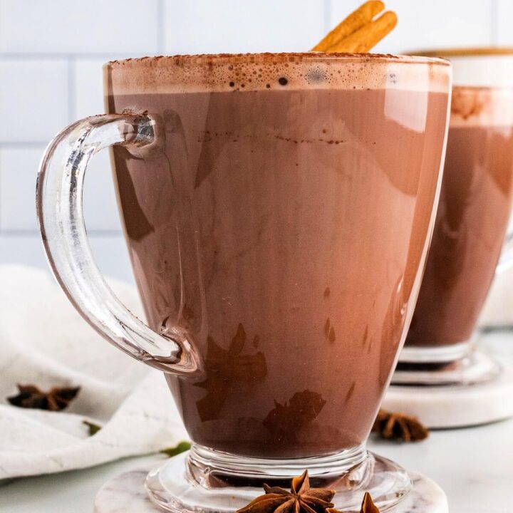 Two glass mugs filled with hot chocolate and garnished with cinnamon sticks and ground cinnamon, sitting on marble coasters with dried star anise pods, cinnamon sticks and cardamom pods sitting around them and a white kitchen towel to the side.