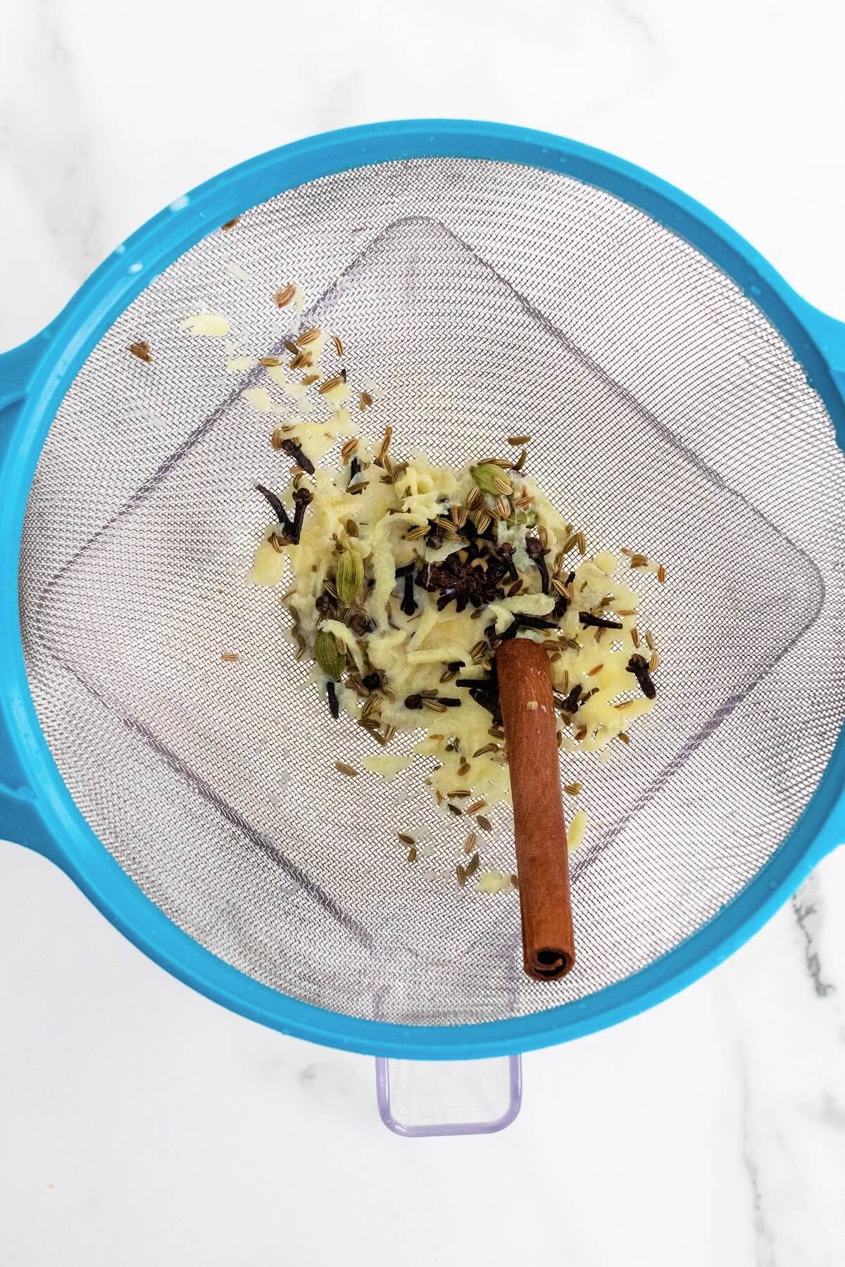 Fine mesh strainer filled with a wet cinnamon stick, whole cloves, grated ginger, fennel seeds and cardamom pods, sitting on top of a blender.