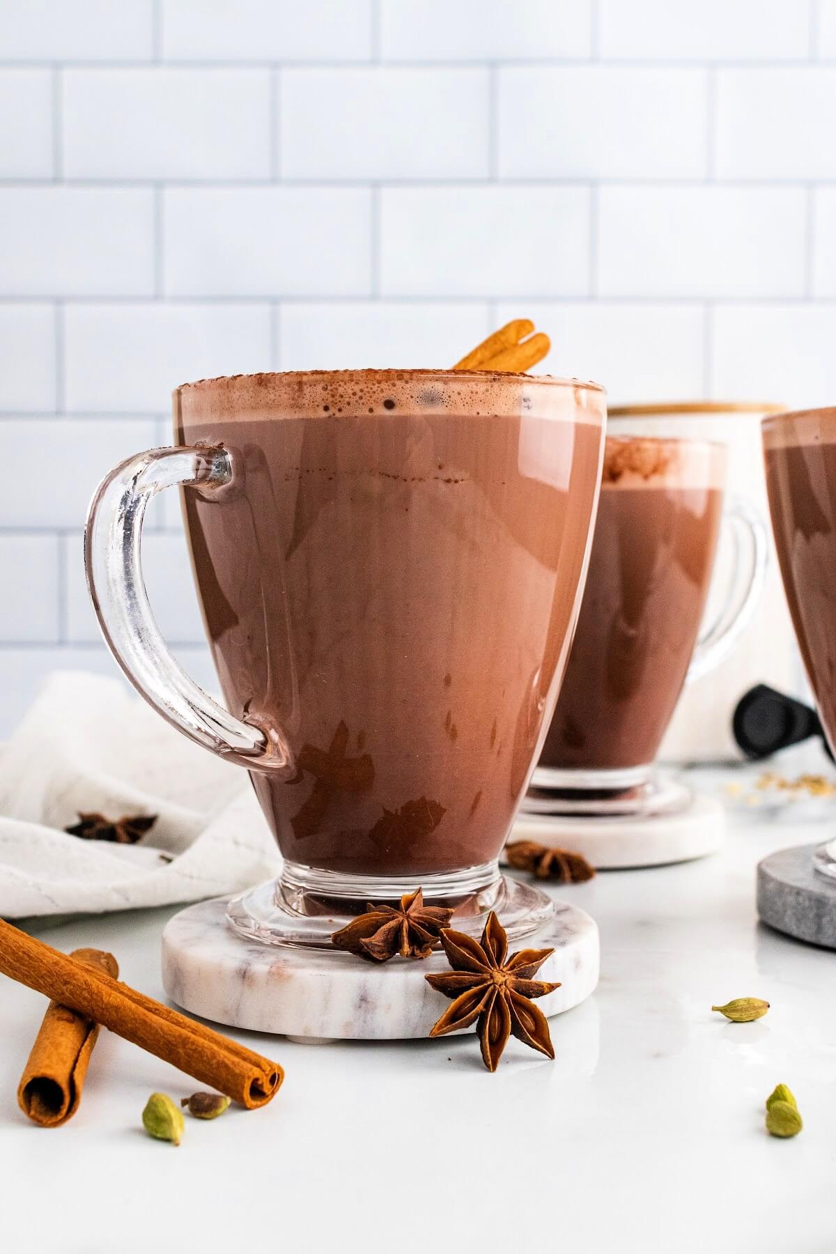 Three glass mugs filled with hot chocolate and garnished with cinnamon sticks and ground cinnamon, sitting on marble coasters with dried star anise pods, cinnamon sticks and cardamom pods sitting around them and a white kitchen towel to the side.