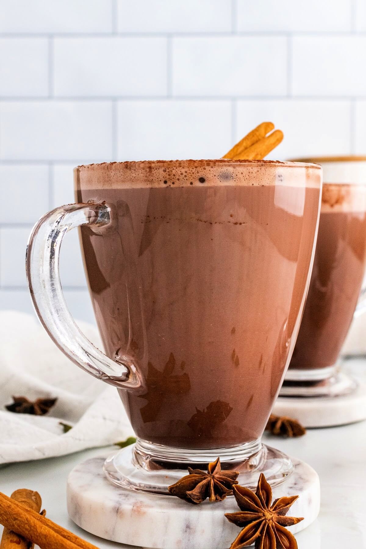 Two glass mugs filled with hot chocolate and garnished with cinnamon sticks and ground cinnamon, sitting on marble coasters with dried star anise pods and cinnamon sticks sitting around them and a white kitchen towel to the side.