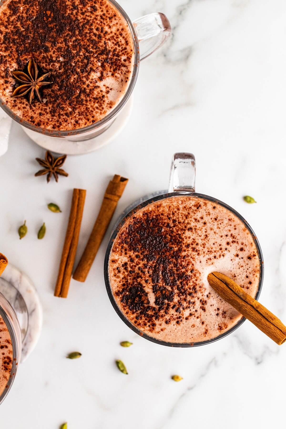 Overhead photo of three glass mugs filled with hot chocolate, one garnished with cinnamon sticks and ground cinnamon and one garnished with ground cinnamon and a star anise pod, sitting on marble coasters with dried star anise pods, cinnamon sticks and cardamom pods sitting around them.
