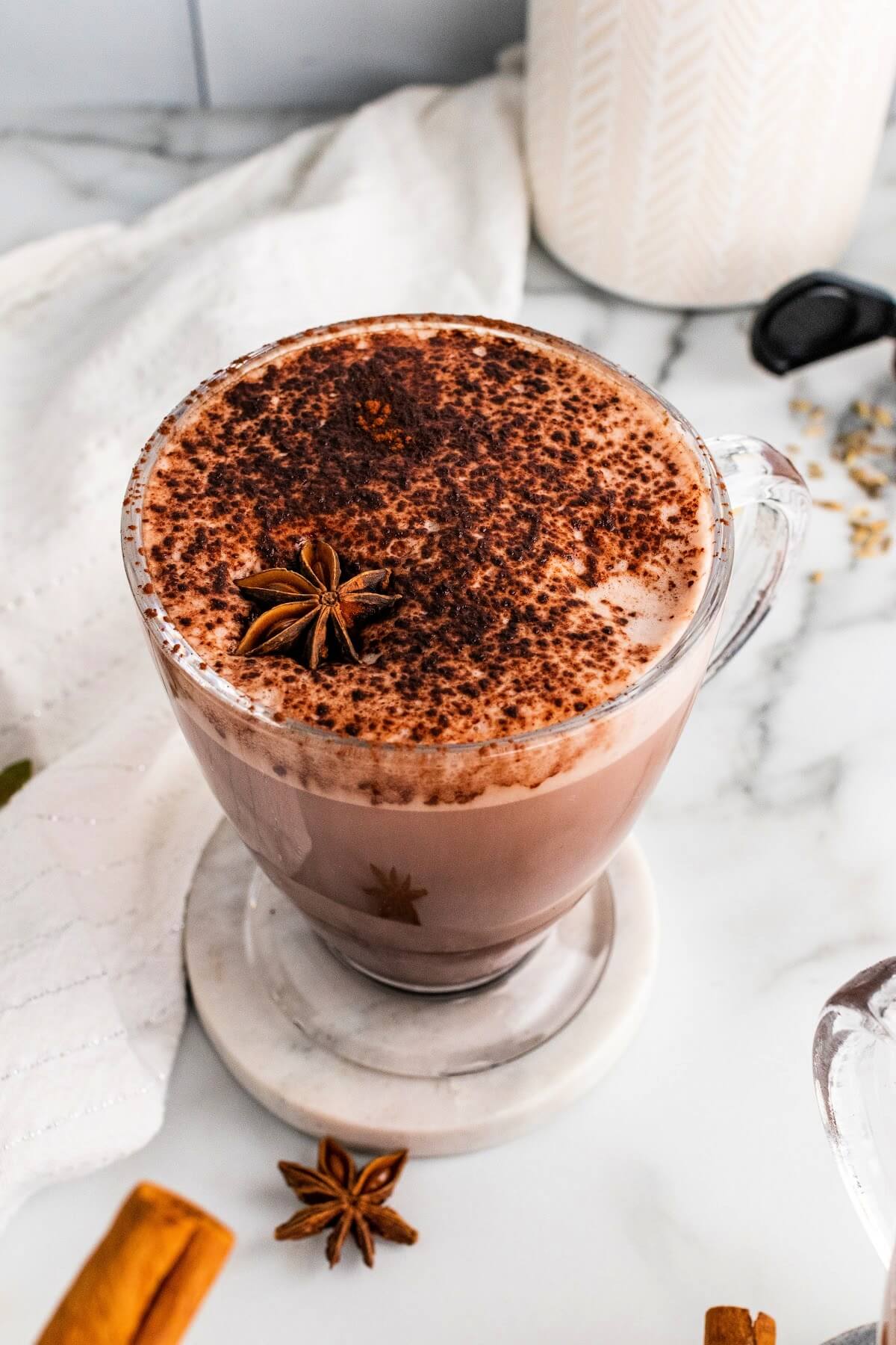 A glass mug filled with hot chocolate and garnished with ground cinnamon and a star anise pod, sitting on a marble coaster with dried star anise pods, cinnamon sticks and fennel seeds sitting around the mug and counter and a white kitchen towel to the side.