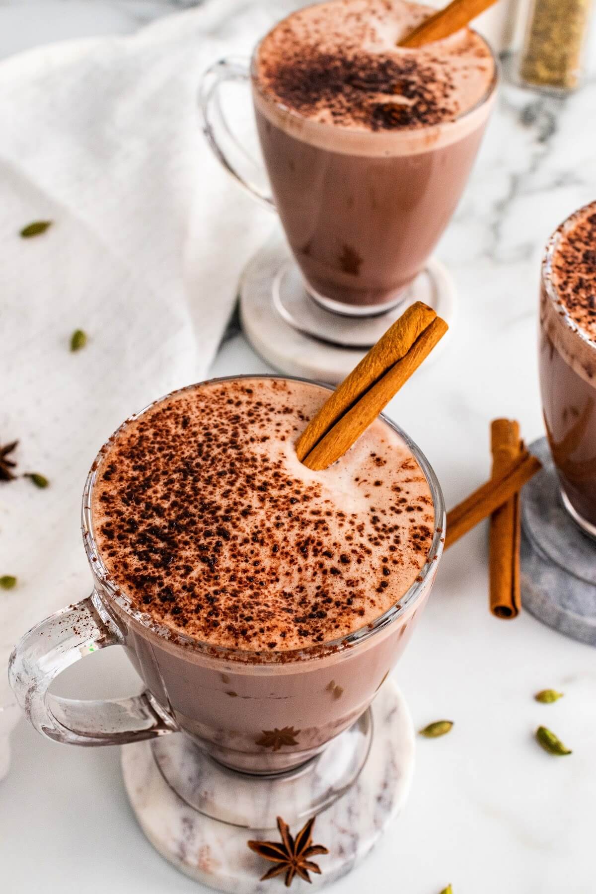 Three glass mugs filled with hot chocolate and garnished with cinnamon sticks and ground cinnamon, sitting on marble coasters with dried star anise pods, cinnamon sticks and cardamom pods sitting around them and a white kitchen towel to the side.