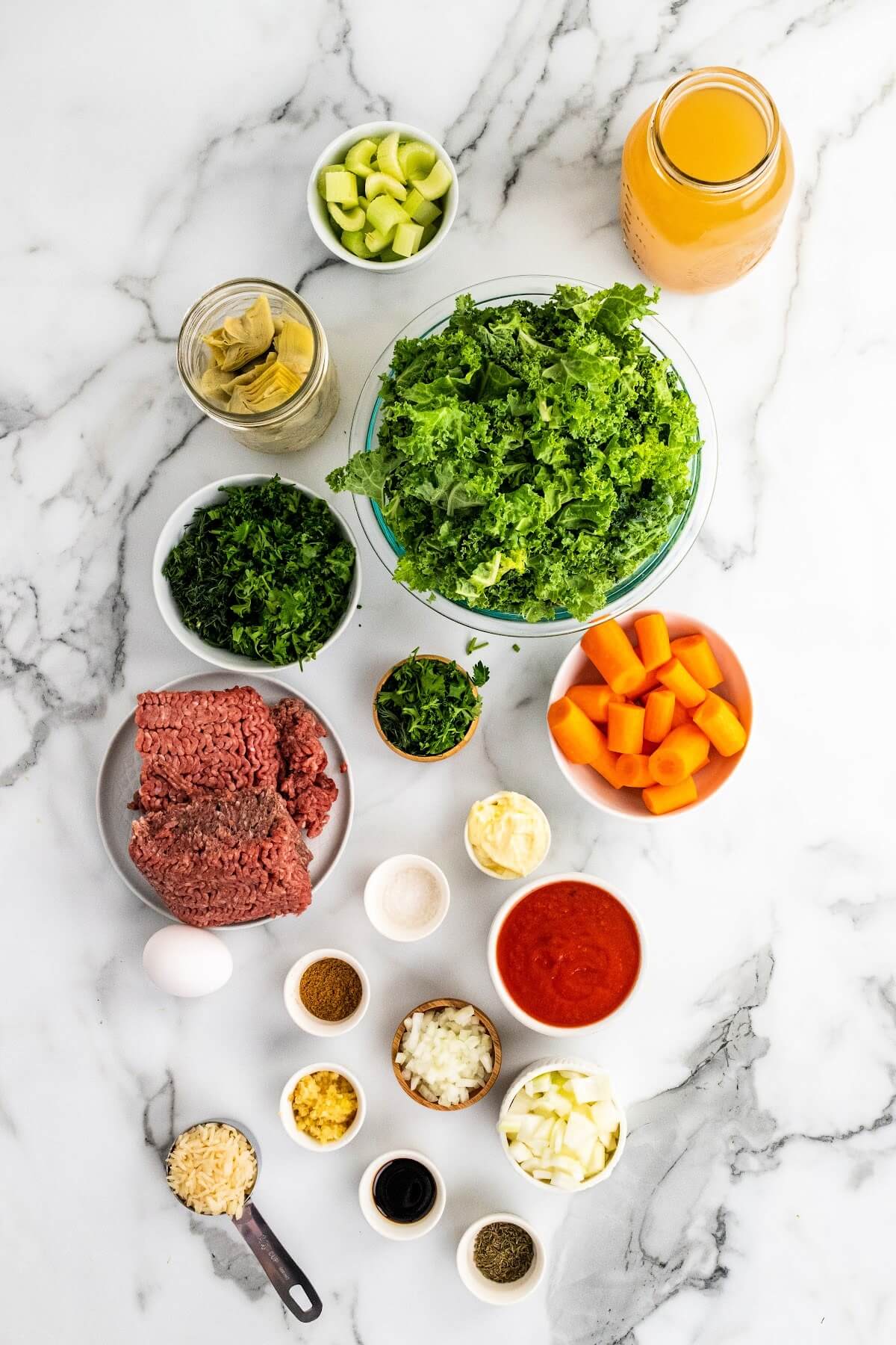 Overhead shot of ingredients - a glass jar with broth, small bowl of chopped celery, glass jar of artichoke hearts, small bowl of chopped herbs, mini bowl of chopped herbs, glass bowl of chopped kale, small bowl of chopped carrots, small bowl of ground beef, 1 egg, small bowl of crushed tomatoes, several mini bowls of spices, chopped garlic and rice.
