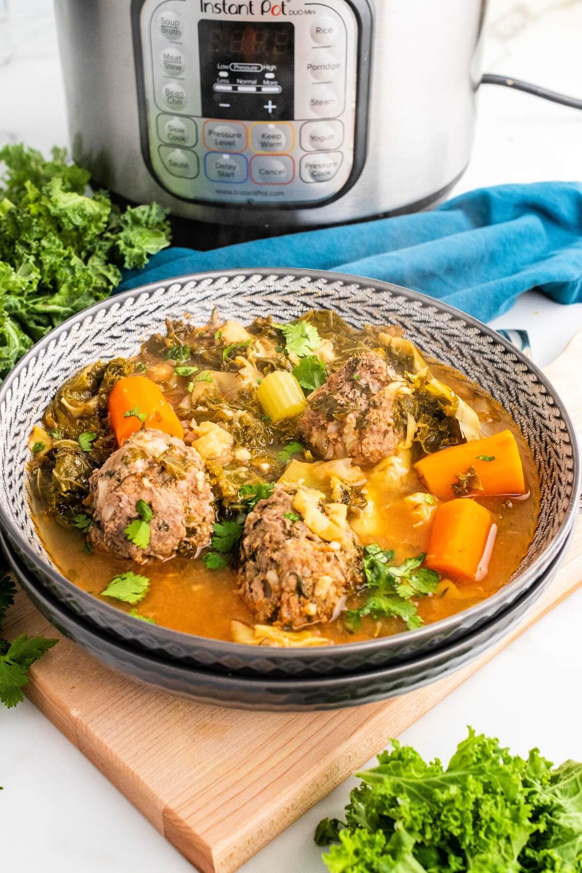 2 stacked bowls with the top bowl full of meatball soup with 3 meatballs, chunks of carrots, vegetables and fresh herbs, sitting on a wood cutting board next to fresh cilantro, fresh kale, a blue cloth napkin and an Instant Pot in the background.