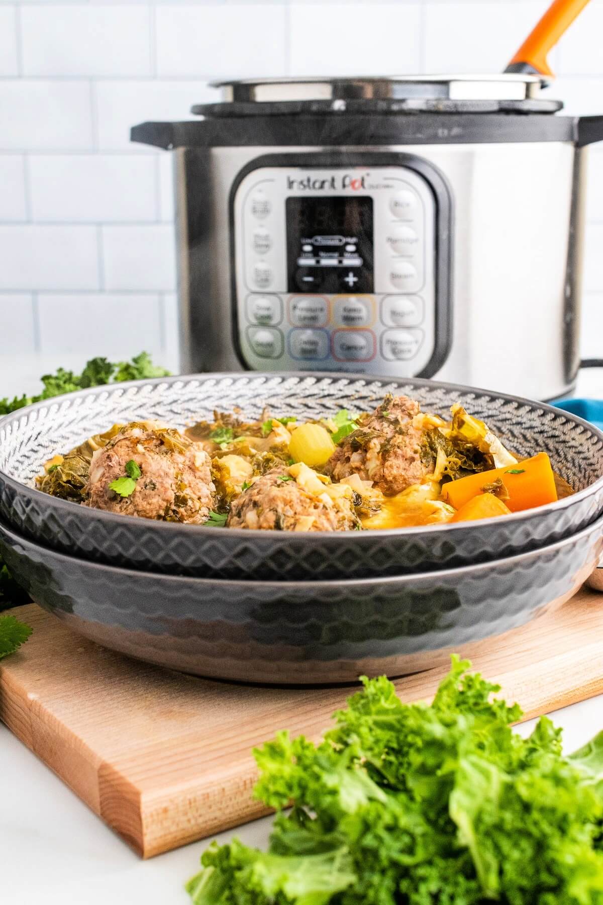 2 stacked bowls with the top bowl full of meatball soup with 3 meatballs, chunks of carrots, vegetables and fresh herbs, sitting on a wood cutting board next to fresh cilantro, fresh kale and an Instant Pot in the background.