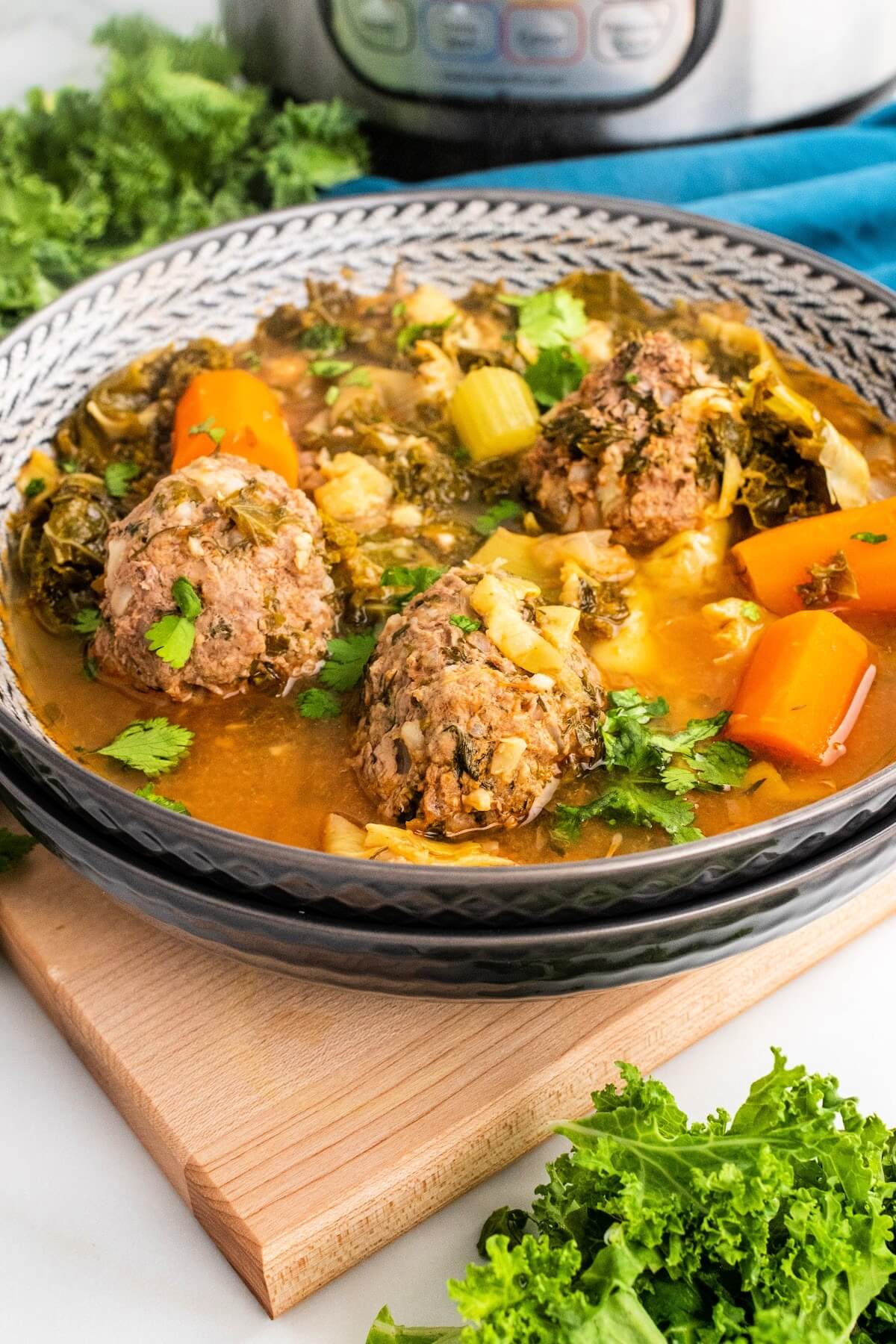 Bowl of meatball soup with 3 meatballs, chunks of carrots, vegetables and fresh herbs, sitting on a wood cutting board next to fresh cilantro, fresh kale and a blue cloth napkin and an Instant Pot in the background.