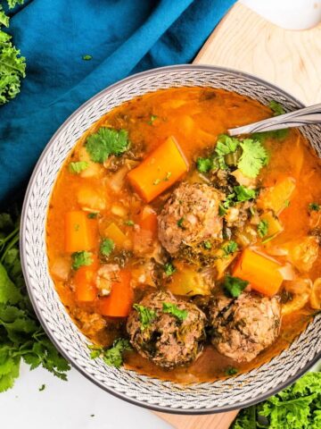 Bowl of meatball soup with 3 meatballs, chunks of carrots, vegetables and fresh herbs, sitting next to fresh cilantro, fresh kale and a blue cloth napkin.