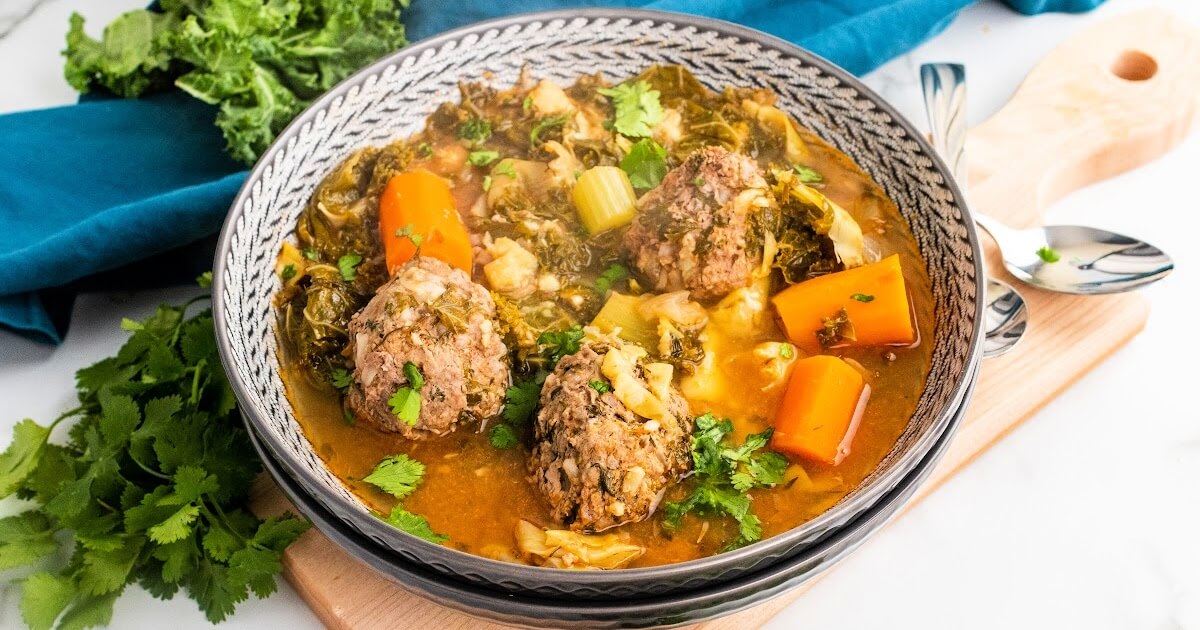 Bowl of meatball soup with 3 meatballs, chunks of carrots, vegetables and fresh herbs, sitting on a wood cutting board next to fresh cilantro, fresh kale and a blue cloth napkin and 2 spoons.