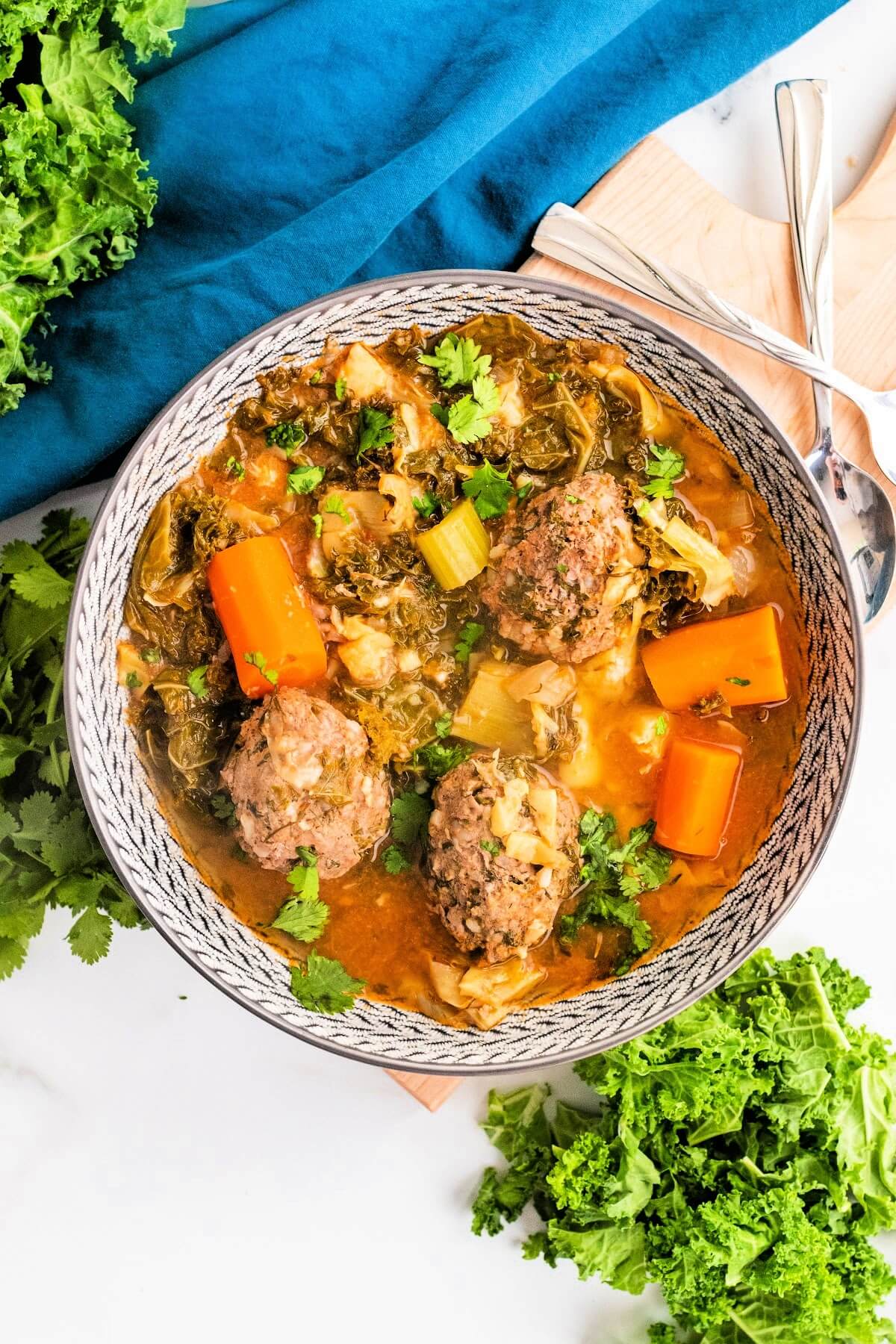 Bowl of meatball soup with 3 meatballs, chunks of carrots, vegetables and fresh herbs, sitting next to fresh cilantro, fresh kale and a blue cloth napkin with 2 spoons.