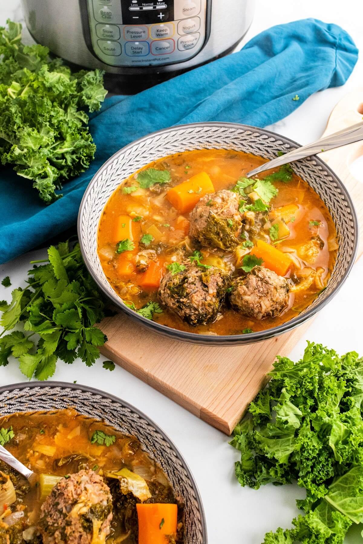 2 bowls of meatball soup with chunks of carrots, vegetables and fresh herbs, sitting next to fresh cilantro, fresh kale and a blue cloth napkin and the Instant Pot in the background.