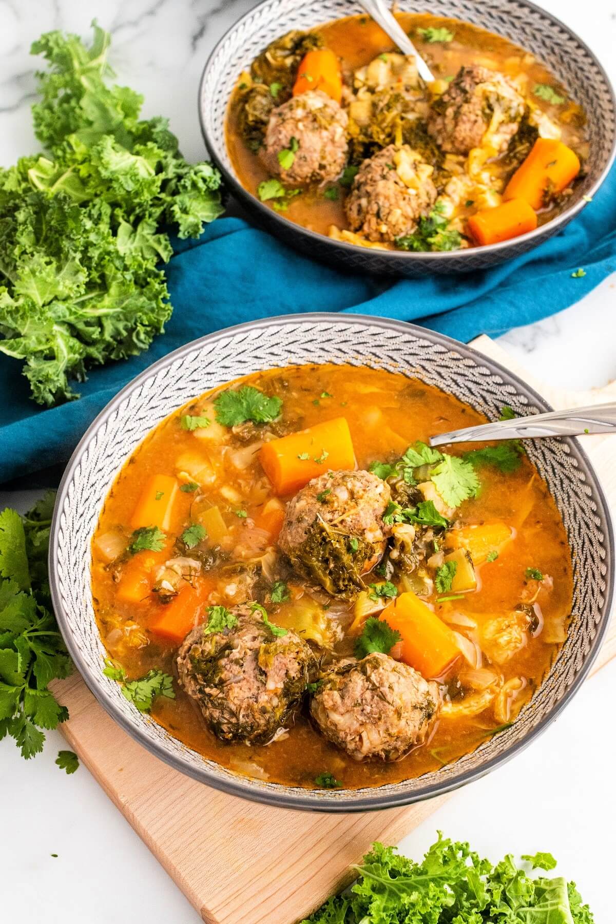 2 bowls of meatball soup with 3 meatballs, chunks of carrots, vegetables and fresh herbs, sitting next to fresh cilantro, fresh kale and a blue cloth napkin.