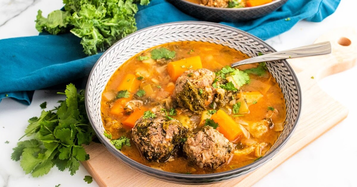 Bowl of meatball soup with 3 meatballs, chunks of carrots, vegetables and fresh herbs with a spoon, sitting on a wood cutting board next to fresh cilantro, fresh kale and a blue cloth napkin with a bowl of soup in the background