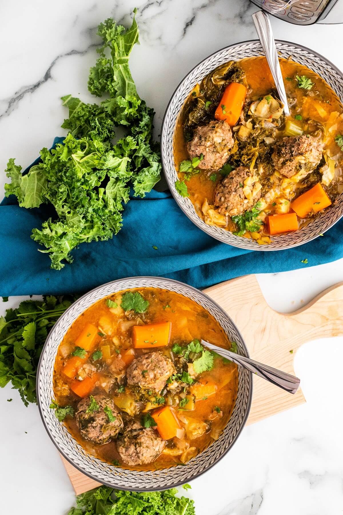 2 bowls of meatball soup with 3 meatballs, chunks of carrots, vegetables and fresh herbs, sitting next to fresh cilantro, fresh kale and a blue cloth napkin.