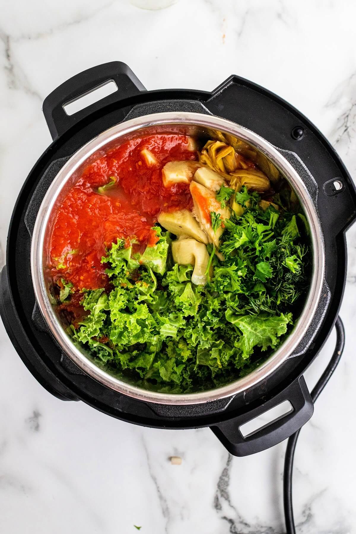 Overhead shot of an Instant Pot filled with crushed tomatoes, fresh kale, fresh herbs and artichoke hearts.