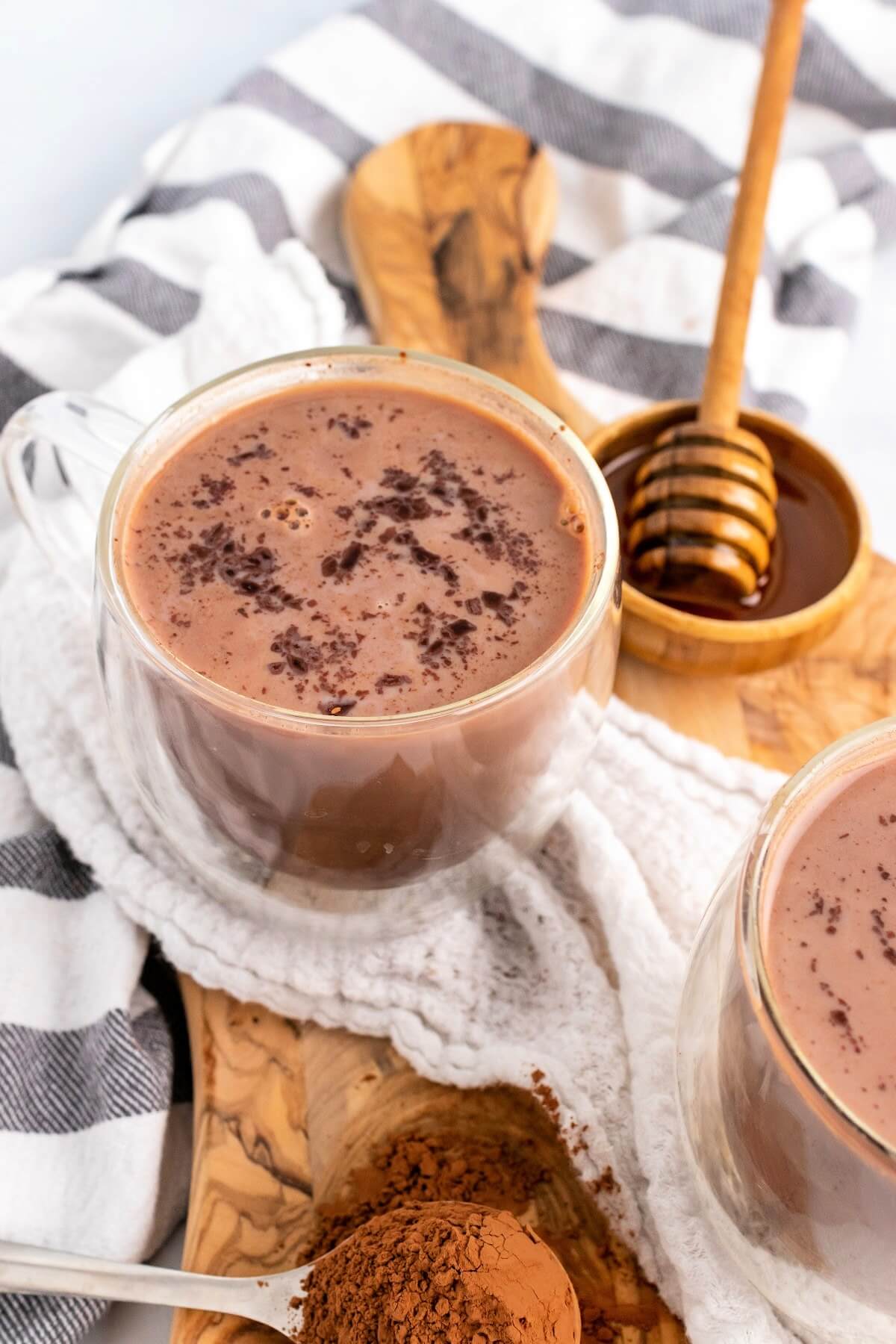 One glass mug and a side view of a glass mug filled with hot chocolate topped with chocolate shavings sitting on a wooden cutting board with a spoon full of cocoa powder and a small bowl full of honey with a honey dipper next to the hot chocolates.