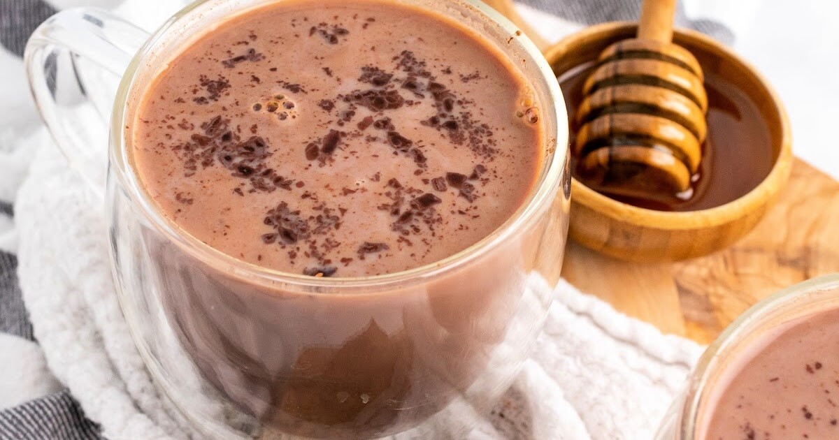 Two glass mugs filled with hot chocolate topped with chocolate shavings sitting on top of a cloth kitchen towel on top of a wooden cutting board next to a small bowl full of honey with a honey dipper next to the hot chocolates.