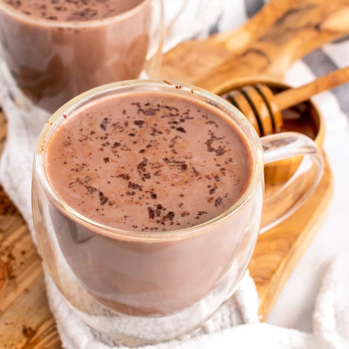 Two glass mugs filled with hot chocolate topped with chocolate shavings sitting on top of a cloth kitchen towel on top of a wooden cutting board next to a small bowl full of honey with a honey dipper next to the hot chocolates.