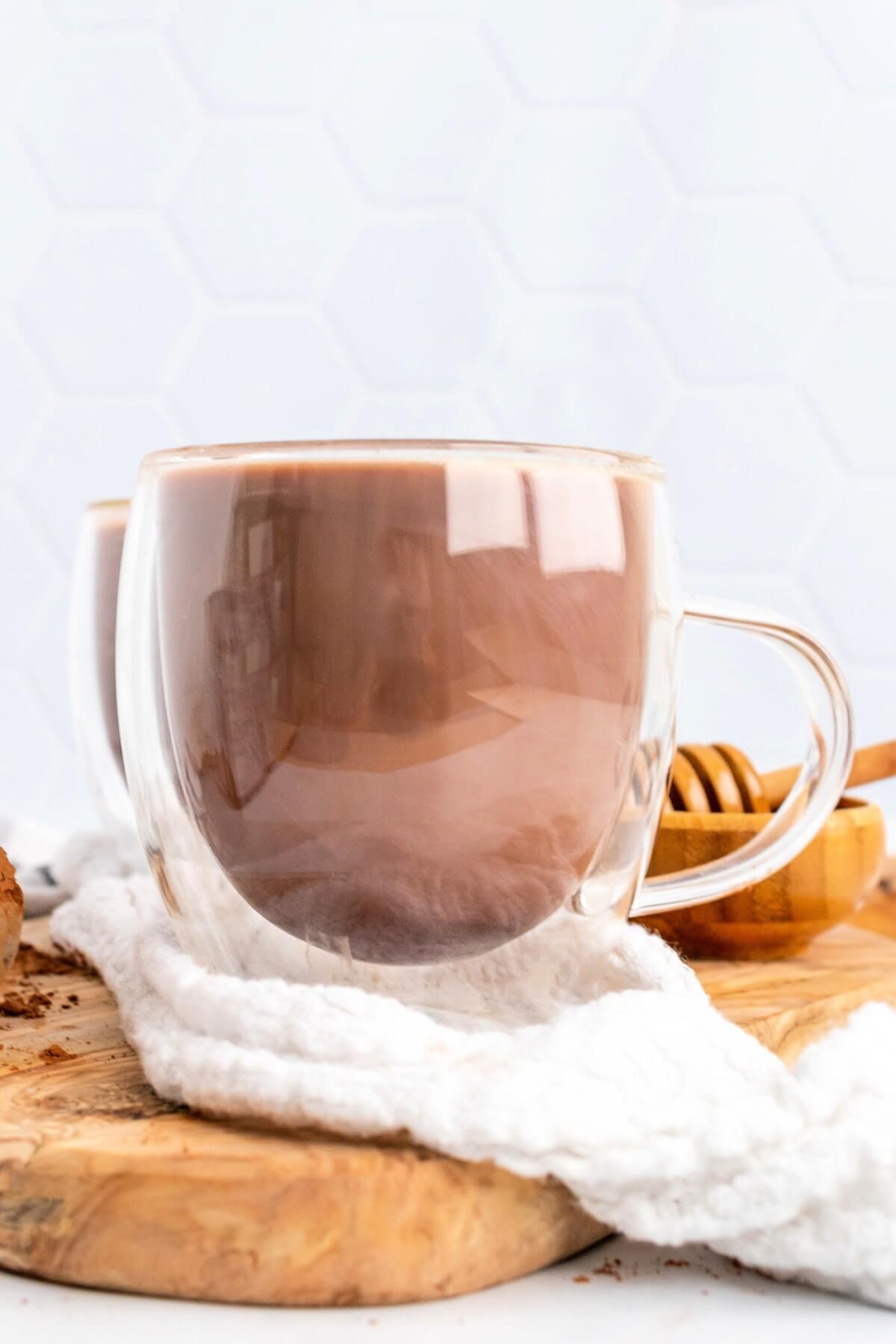 Glass mug filled with hot chocolate sitting on a wooden cutting board and a cloth kitchen towel with a honey dipper sitting behind it.