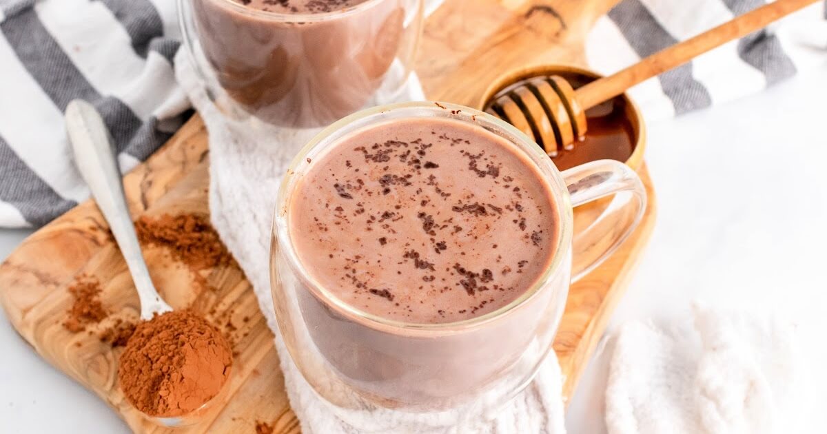 Two glass mugs filled with hot chocolate topped with chocolate shavings sitting on top of a cloth kitchen towel on top of a wooden cutting board next to a spoon full of cocoa powder and a small bowl full of honey with a honey dipper next to the hot chocolates.