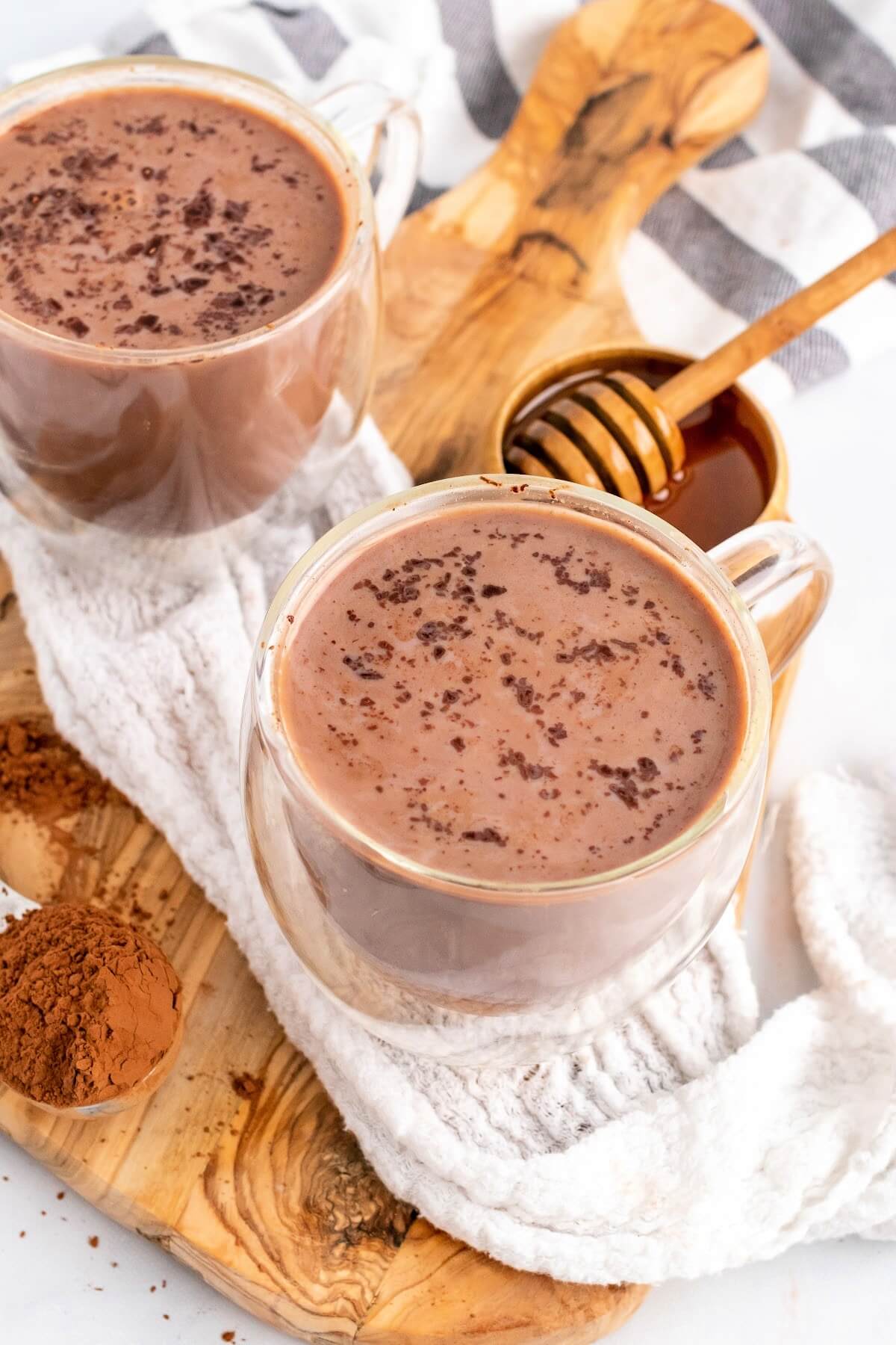 Two glass mugs filled with hot chocolate topped with chocolate shavings sitting on top of a cloth kitchen towel on top of a wooden cutting board next to a spoon full of cocoa powder and a small bowl full of honey with a honey dipper next to the hot chocolates.