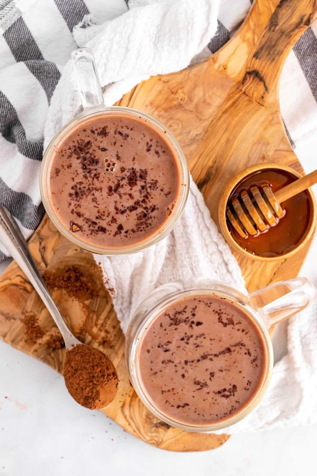 Two glass mugs filled with hot chocolate and topped with chocolate shavings sitting on a wooden cutting board with a spoon full of cocoa powder and a small bowl full of honey with a honey dipper next to the hot chocolates.