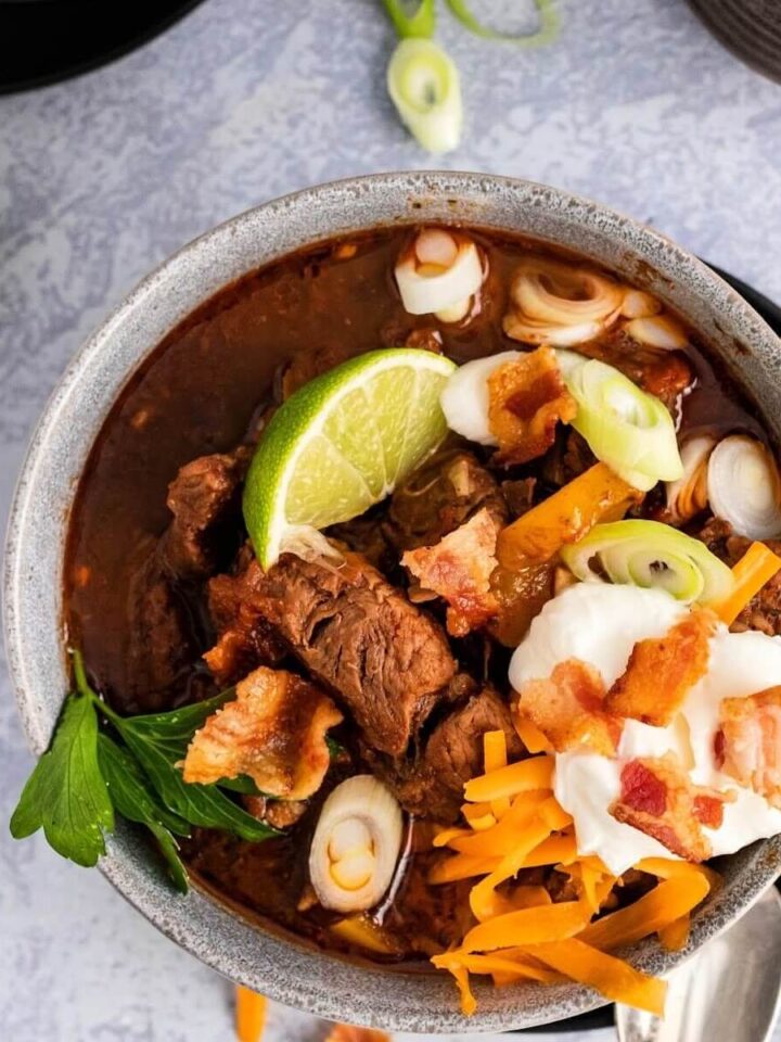 Bowl full of meat chili with chunks of stew meat, topped with shredded cheddar cheese, sour cream, bacon pieces, green onions, fresh cilantro and a lime wedge, sitting on a plate with a spoon next to it and sliced green onions and a lime wedge in the background.