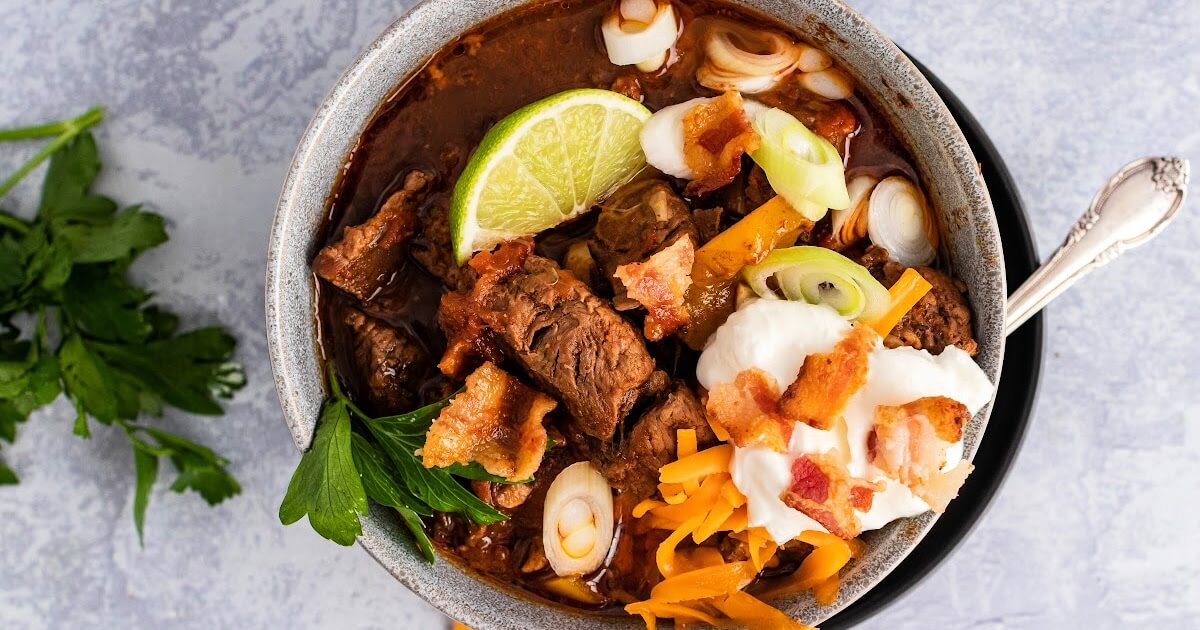 Bowl full of meat chili with chunks of stew meat, topped with shredded cheddar cheese, sour cream, bacon pieces, green onions, fresh cilantro and a lime wedge, sitting on a plate with a spoon on the plate and fresh cilantro sprigs next to the bowl of chili.